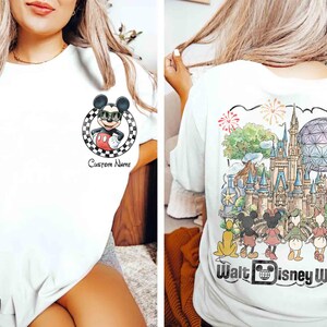 May include: White t-shirt featuring Mickey Mouse wearing sunglasses, a red shorts, and a black and white checkered circle. The back of the shirt displays a colorful illustration of Walt Disney World with characters and fireworks.