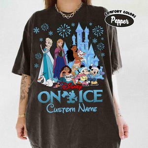 Puede incluir: Camiseta gris oscuro con personajes de Disney de Frozen, Moana y Mickey y Minnie Mouse. La camiseta tiene el texto "Disney On Ice" y "Custom Name" en azul. La camiseta tambi&eacute;n tiene las palabras "Comfort Colors Pepper" en un &oacute;valo blanco.