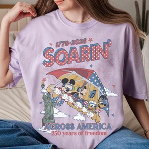 May include: A lilac-colored t-shirt featuring a graphic of Mickey Mouse, Goofy, and Donald Duck flying under a red, white, and blue umbrella. The text "SOARIN'" and "ACROSS AMERICA" are displayed above and below the graphic, with the years "1776-2026" and "250 years of freedom".
