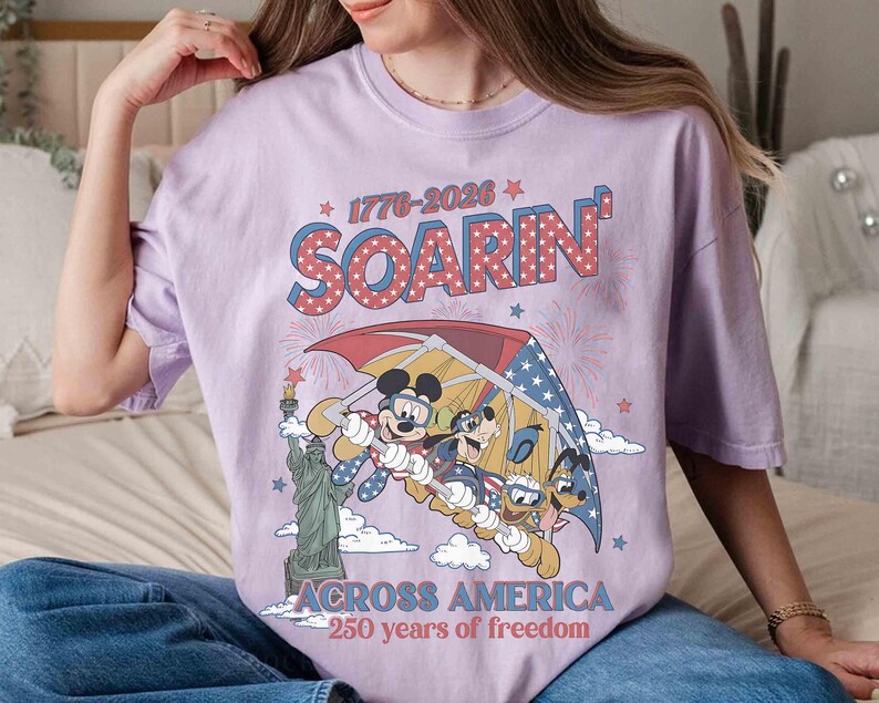 May include: A lilac-colored t-shirt featuring a graphic of Mickey Mouse, Goofy, and Donald Duck flying under a red, white, and blue umbrella. The text "SOARIN'" and "ACROSS AMERICA" are displayed above and below the graphic, with the years "1776-2026" and "250 years of freedom".