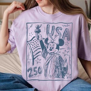 K&ouml;nnte beinhalten: Helllila T-Shirt mit einer Skizze von Minnie Mouse als Freiheitsstatue, die eine Fackel und die amerikanische Flagge h&auml;lt. Das Wort "USA" steht &uuml;ber Minnie, und die Zahl "250" steht darunter.