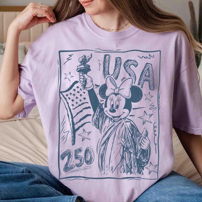 K&ouml;nnte beinhalten: Helllila T-Shirt mit einer Skizze von Minnie Mouse als Freiheitsstatue, die eine Fackel und die amerikanische Flagge h&auml;lt. Das Wort "USA" steht &uuml;ber Minnie, und die Zahl "250" steht darunter.