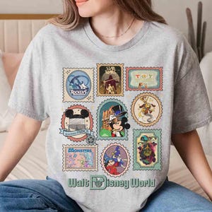 Puede incluir: Una camiseta gris con un collage de coloridos dise&ntilde;os de sellos postales con varios personajes de dibujos animados y logotipos. Los sellos incluyen "Rockers", "Star Wars", "Toy Story" y "Monsters". El texto "Walt Disney World" est&aacute; impreso en la parte inferior.