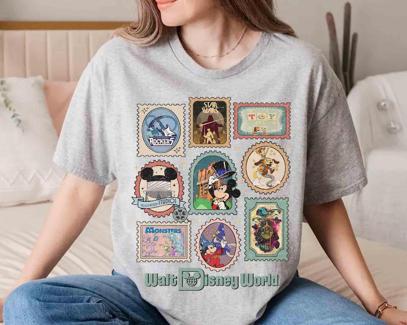 Puede incluir: Una camiseta gris con un collage de coloridos dise&ntilde;os de sellos postales con varios personajes de dibujos animados y logotipos. Los sellos incluyen "Rockers", "Star Wars", "Toy Story" y "Monsters". El texto "Walt Disney World" est&aacute; impreso en la parte inferior.
