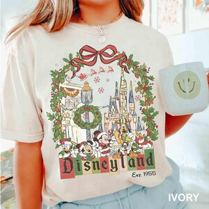 Op de afbeelding: Ivoor kleurig T-shirt met een Disneyland kerstontwerp. De afbeelding toont een krans, kasteel en Disney-figuren. Het woord "Disneyland" staat in een feestelijk lettertype, met "Est. 1955" eronder.