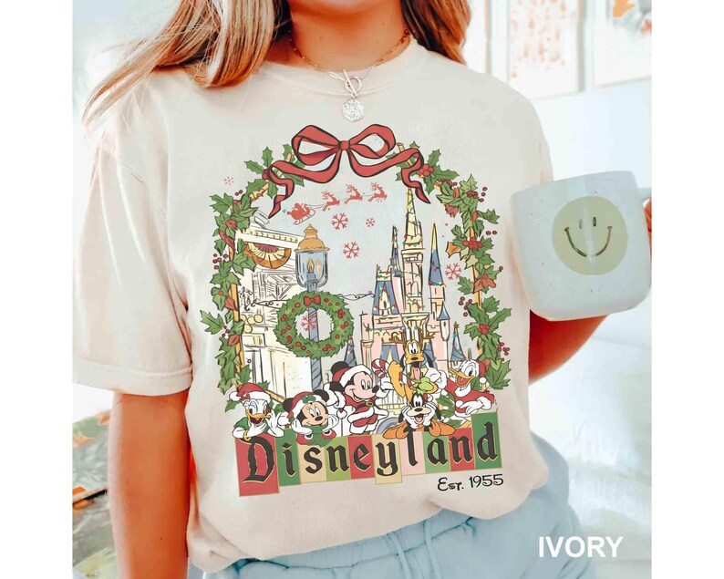 Op de afbeelding: Ivoor kleurig T-shirt met een Disneyland kerstontwerp. De afbeelding toont een krans, kasteel en Disney-figuren. Het woord "Disneyland" staat in een feestelijk lettertype, met "Est. 1955" eronder.