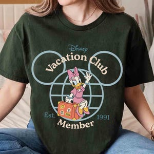 Puede incluir: Camiseta verde oscuro con un dise&ntilde;o de Disney Vacation Club. El gr&aacute;fico incluye a Daisy Duck con un vestido rosa y una maleta, dentro de un contorno de la cabeza de Mickey Mouse. El texto incluye "Vacation Club", "Member" y "Est. 1991".