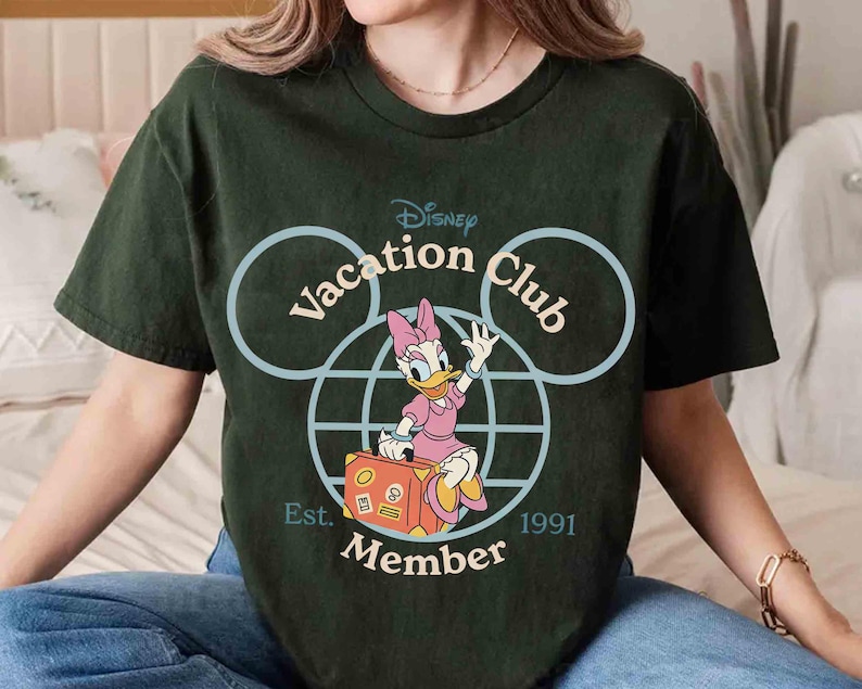 Puede incluir: Camiseta verde oscuro con un dise&ntilde;o de Disney Vacation Club. El gr&aacute;fico incluye a Daisy Duck con un vestido rosa y una maleta, dentro de un contorno de la cabeza de Mickey Mouse. El texto incluye "Vacation Club", "Member" y "Est. 1991".