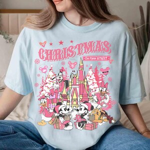 May include: A light blue t-shirt featuring a pink graphic with the word "CHRISTMAS" above a castle and cartoon characters. The graphic includes Mickey and Minnie Mouse, Donald Duck, and other characters, with Christmas trees and snowflakes.