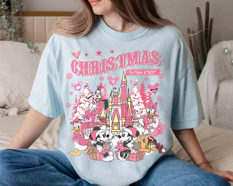 May include: A light blue t-shirt featuring a pink graphic with the word "CHRISTMAS" above a castle and cartoon characters. The graphic includes Mickey and Minnie Mouse, Donald Duck, and other characters, with Christmas trees and snowflakes.