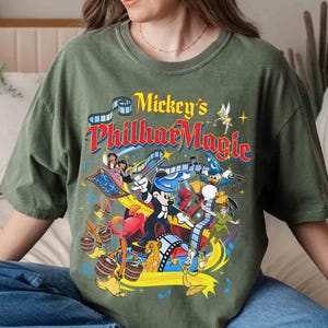 K&ouml;nnte beinhalten: Ein olivgr&uuml;nes T-Shirt mit dem Text "Mickey's PhilharMagic" in einem farbenfrohen Vintage-Design. Die Grafik zeigt verschiedene Disney-Figuren wie Mickey Mouse, Donald Duck und Peter Pan, mit einem Filmstreifen-Motiv.