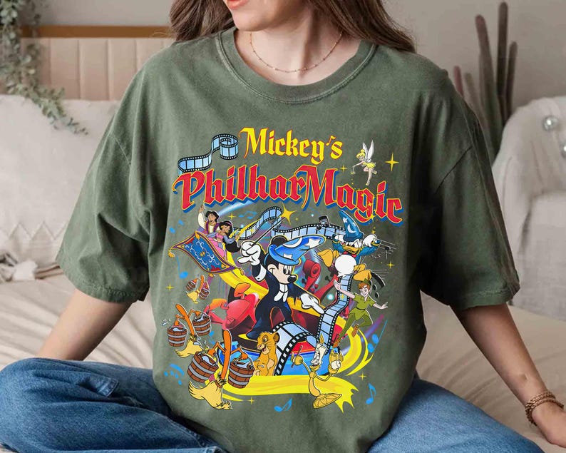 K&ouml;nnte beinhalten: Ein olivgr&uuml;nes T-Shirt mit dem Text "Mickey's PhilharMagic" in einem farbenfrohen Vintage-Design. Die Grafik zeigt verschiedene Disney-Figuren wie Mickey Mouse, Donald Duck und Peter Pan, mit einem Filmstreifen-Motiv.