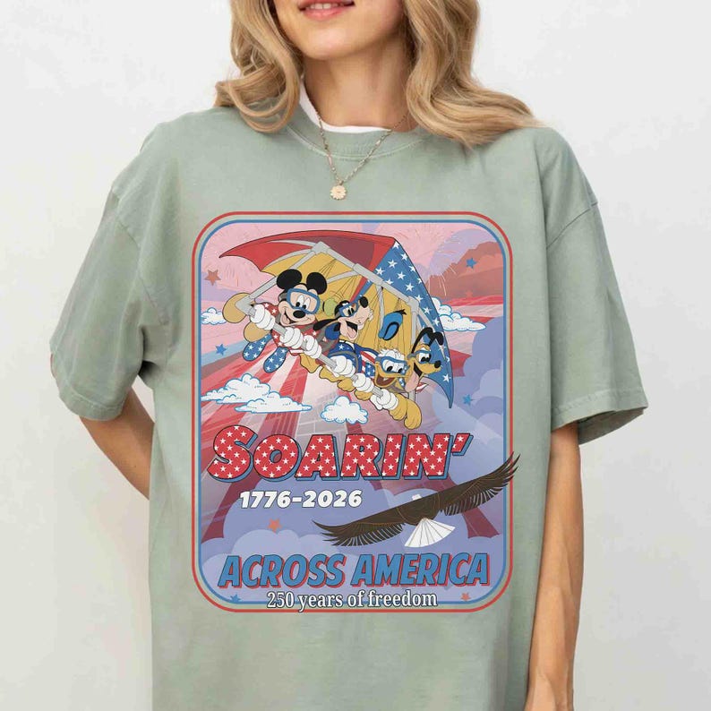 May include: Sage green t-shirt featuring a colorful graphic of Mickey Mouse, Donald Duck, and Goofy soaring through the sky. The design includes the text "Soarin' Across America" and "1776-2026", celebrating 250 years of freedom.