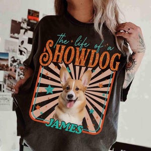 May include: Dark gray t-shirt featuring a smiling corgi dog graphic with the text "the life of a SHOWDOG" and the name "JAMES". The design includes a retro-style background with stars and stripes.