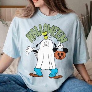 May include: Light blue Halloween-themed t-shirt featuring a cartoon character dressed as a ghost, holding a Mickey Mouse pumpkin bucket. The word "HALLOWEEN" is printed above the character in green. The character is wearing blue pants and brown shoes.