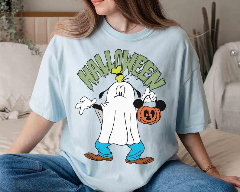 May include: Light blue Halloween-themed t-shirt featuring a cartoon character dressed as a ghost, holding a Mickey Mouse pumpkin bucket. The word "HALLOWEEN" is printed above the character in green. The character is wearing blue pants and brown shoes.