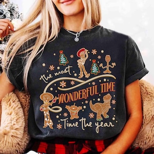 Puede incluir: Camiseta gris oscuro con personajes de Toy Story estilo pan de jengibre, incluyendo a Woody y Jessie, con el texto "The most wonderful time of the year". El dise&ntilde;o incluye copos de nieve y &aacute;rboles de Navidad.