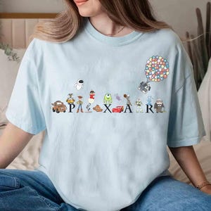 K&ouml;nnte beinhalten: Hellblaues T-Shirt mit dem Wort "PIXAR" und Illustrationen von Zeichentrickfiguren. Das Design umfasst Charaktere aus verschiedenen Pixar-Filmen wie "Cars", "Toy Story" und "Monster AG". Ein bunter Balloncluster ist ebenfalls abgebildet.