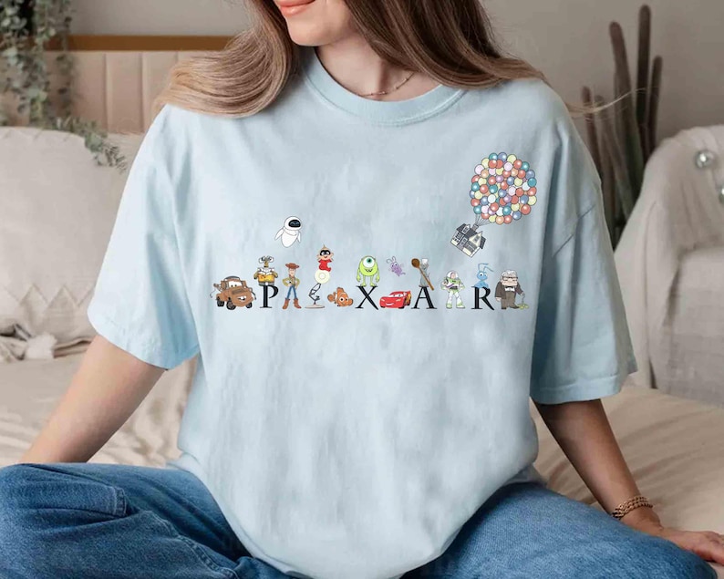 K&ouml;nnte beinhalten: Hellblaues T-Shirt mit dem Wort "PIXAR" und Illustrationen von Zeichentrickfiguren. Das Design umfasst Charaktere aus verschiedenen Pixar-Filmen wie "Cars", "Toy Story" und "Monster AG". Ein bunter Balloncluster ist ebenfalls abgebildet.