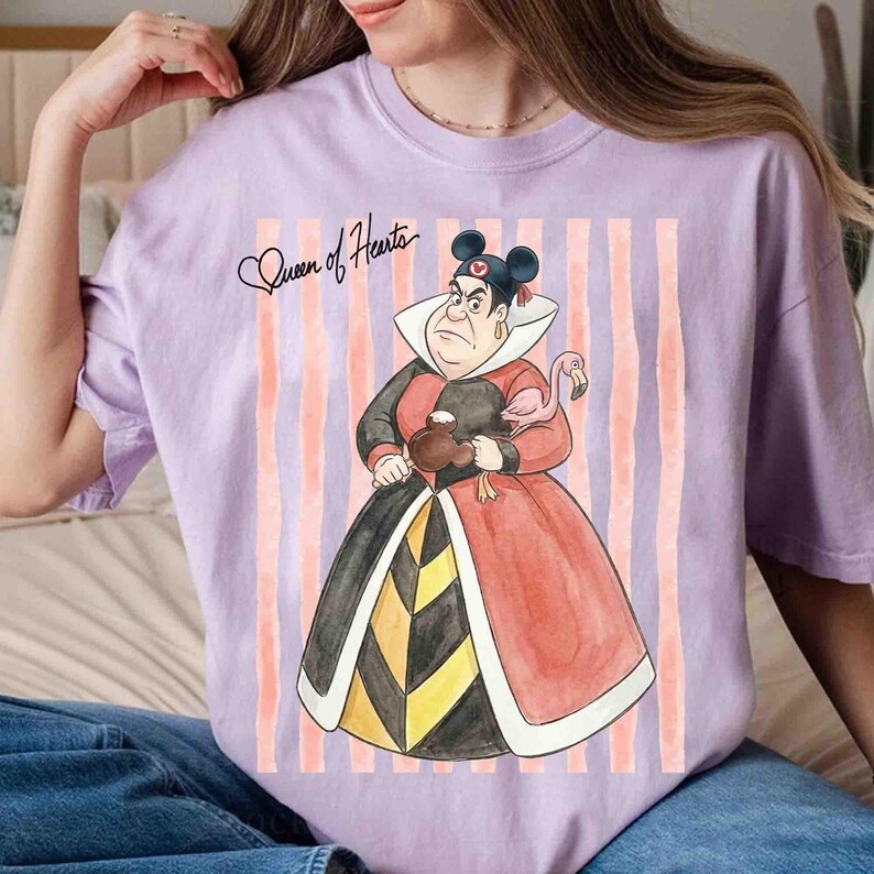 May include: A light purple t-shirt featuring a graphic of the Queen of Hearts wearing Mickey Mouse ears, holding a brown Mickey-shaped cookie and a pink flamingo. The text "Queen of Hearts" is written in black script above the character.