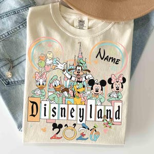 May include: Cream-colored Disneyland 2020 sweatshirt featuring Mickey Mouse, Minnie Mouse, Goofy, Donald Duck, and Daisy Duck. The design includes a castle, fireworks, and the word "Disneyland" in a block letter style.