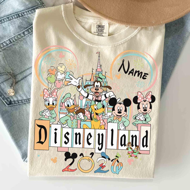 May include: Cream-colored Disneyland 2020 sweatshirt featuring Mickey Mouse, Minnie Mouse, Goofy, Donald Duck, and Daisy Duck. The design includes a castle, fireworks, and the word "Disneyland" in a block letter style.