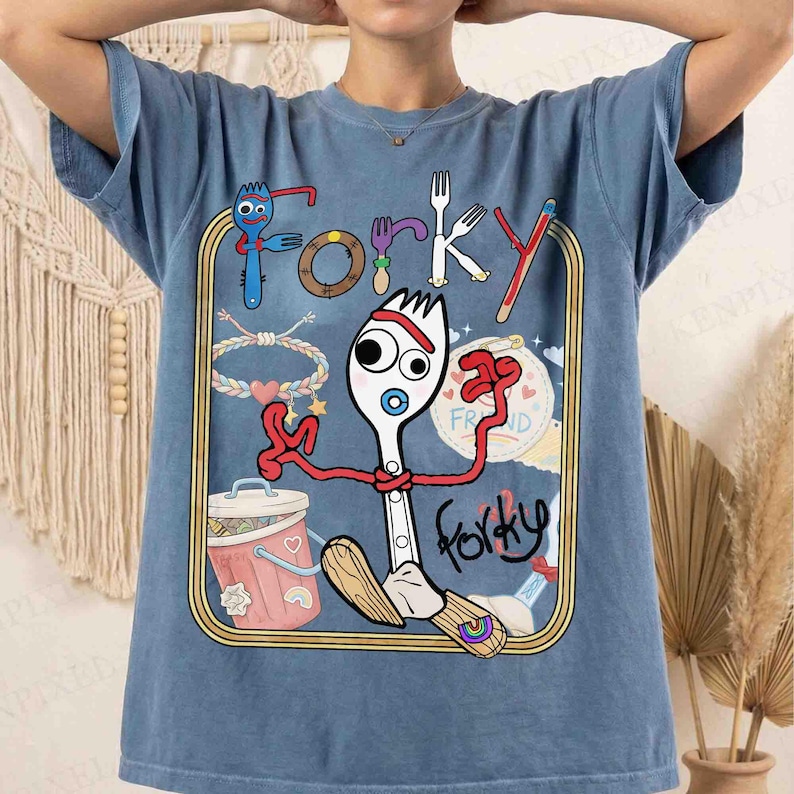 May include: A blue t-shirt featuring a graphic print of the character Forky from Toy Story. The design includes the text Forky, a pink trash can, a braided bracelet, and various utensils inside a gold-colored rectangular frame on the front.