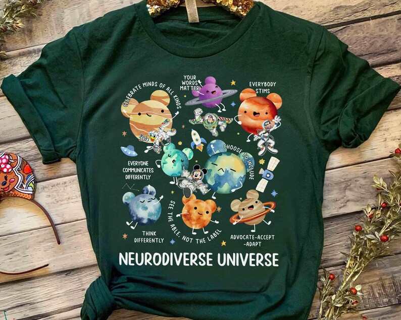 Op de afbeelding: Groen T-shirt met een afbeelding van planeten en sterren. De tekst "Celebrate Minds of All Kinds" en "Neurodiverse Universe" staat op het T-shirt gedrukt.