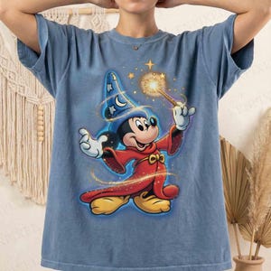 May include: Blue t-shirt featuring a graphic of Mickey Mouse dressed as a wizard. Mickey is wearing a red robe and a blue wizard hat with stars and moons. He is holding a wand with a glowing star at the end.