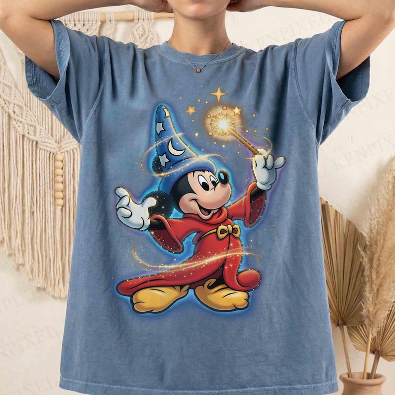 May include: Blue t-shirt featuring a graphic of Mickey Mouse dressed as a wizard. Mickey is wearing a red robe and a blue wizard hat with stars and moons. He is holding a wand with a glowing star at the end.