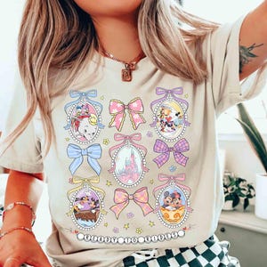 May include: A light beige t-shirt with a graphic print of cartoon characters and bows. The characters are in pastel colors and are surrounded by bows in pink, blue, and yellow. The text "Believe in Magic" is printed at the bottom of the shirt.