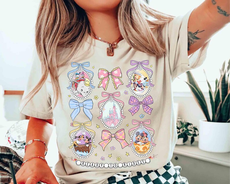 May include: A light beige t-shirt with a graphic print of cartoon characters and bows. The characters are in pastel colors and are surrounded by bows in pink, blue, and yellow. The text "Believe in Magic" is printed at the bottom of the shirt.