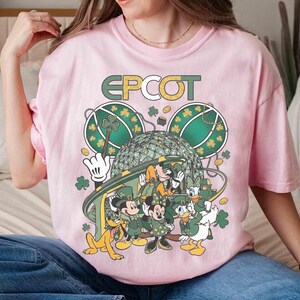 May include: A light pink t-shirt featuring a graphic of Disney characters dressed in green for St. Patrick's Day. The word "EPCOT" is printed above the characters. The design includes Mickey Mouse, Minnie Mouse, Donald Duck, and Pluto.