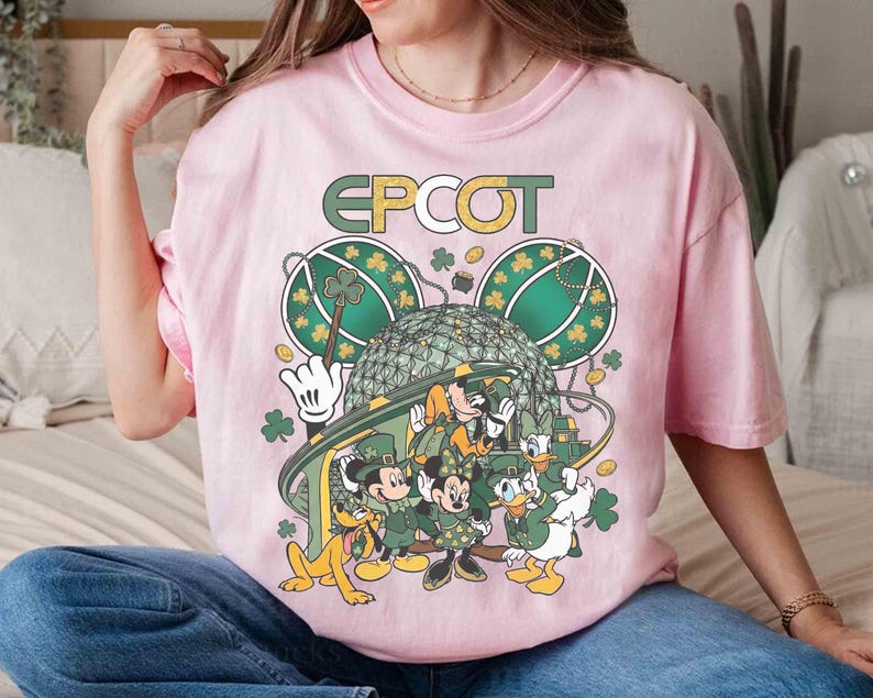 May include: A light pink t-shirt featuring a graphic of Disney characters dressed in green for St. Patrick's Day. The word "EPCOT" is printed above the characters. The design includes Mickey Mouse, Minnie Mouse, Donald Duck, and Pluto.