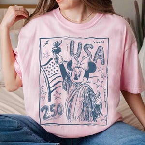K&ouml;nnte beinhalten: Hellrosa T-Shirt mit einer dunkelblauen Grafik von Minnie Mouse als Freiheitsstatue, die eine Fackel und die amerikanische Flagge h&auml;lt. Das Wort "USA" steht &uuml;ber Minnie, und die Zahl "250" steht darunter. Das Design ist dunkelblau umrandet.
