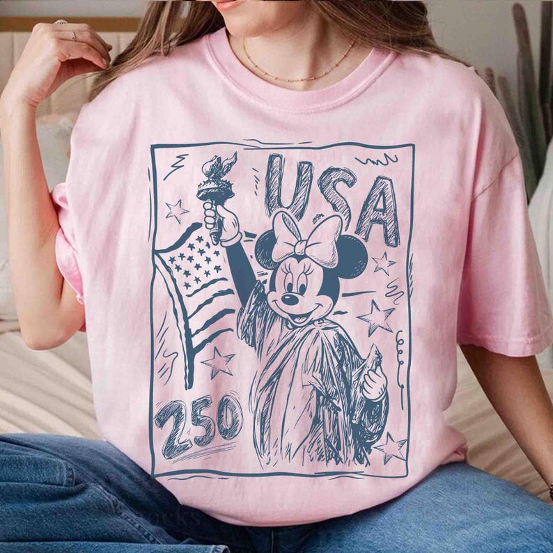 K&ouml;nnte beinhalten: Hellrosa T-Shirt mit einer dunkelblauen Grafik von Minnie Mouse als Freiheitsstatue, die eine Fackel und die amerikanische Flagge h&auml;lt. Das Wort "USA" steht &uuml;ber Minnie, und die Zahl "250" steht darunter. Das Design ist dunkelblau umrandet.