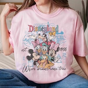 May include: Pink t-shirt featuring a Disneyland graphic with Mickey Mouse, Minnie Mouse, Donald Duck, Daisy Duck, Goofy, and Pluto. The text reads "Disneyland est. 1955 Where dreams come true."