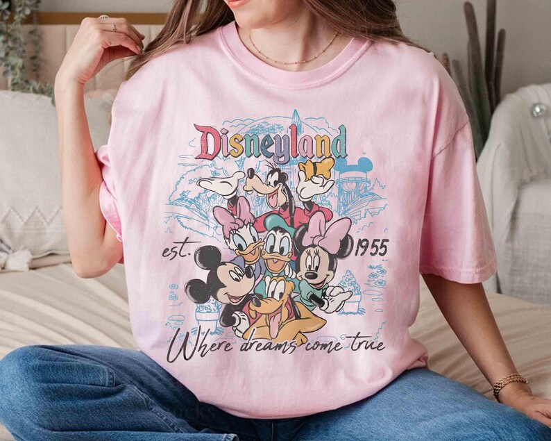 May include: Pink t-shirt featuring a Disneyland graphic with Mickey Mouse, Minnie Mouse, Donald Duck, Daisy Duck, Goofy, and Pluto. The text reads "Disneyland est. 1955 Where dreams come true."