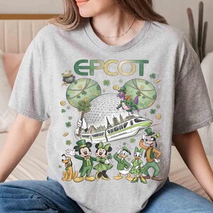 May include: A grey t-shirt with a St. Patrick's Day theme, displaying the word "EPCOT" in green and gold. The design includes Mickey Mouse, Minnie Mouse, Donald Duck, Daisy Duck, Goofy, and Pluto in green attire, alongside a monorail and Spaceship Earth.