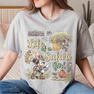 May include: A gray t-shirt featuring a colorful graphic design with the text "Life of a Safari Girl." The design includes cartoon characters, a map, and the words "Disney's Animal Kingdom."