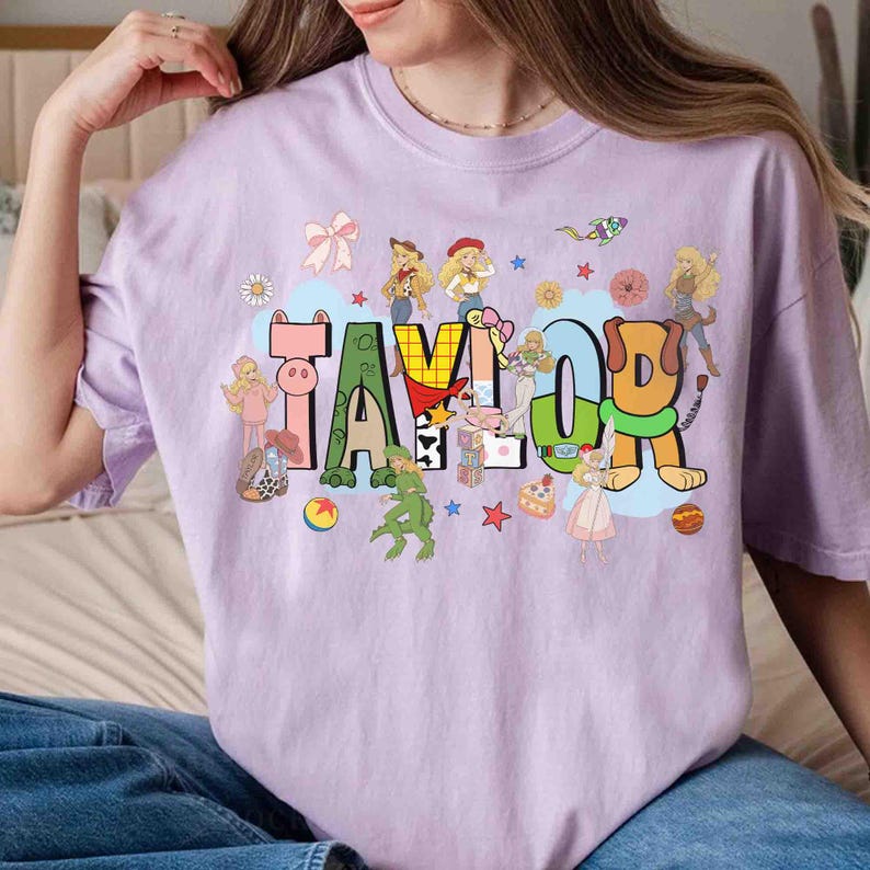 May include: A light purple t-shirt featuring the name TAYLOR in large, colorful, block-style letters. The design is decorated with various cartoon characters, stars, flowers, a bow, and small toys, creating a playful and vibrant graphic print.
