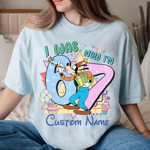 May include: Light blue t-shirt featuring a cartoon character holding a gift and a party hat. The shirt has the text "I WAS 6, NOW I'M 7" with a customizable name option. Colorful birthday-themed graphics include balloons, a cake, and a cupcake.