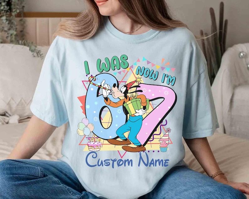 May include: Light blue t-shirt featuring a cartoon character holding a gift and a party hat. The shirt has the text "I WAS 6, NOW I'M 7" with a customizable name option. Colorful birthday-themed graphics include balloons, a cake, and a cupcake.