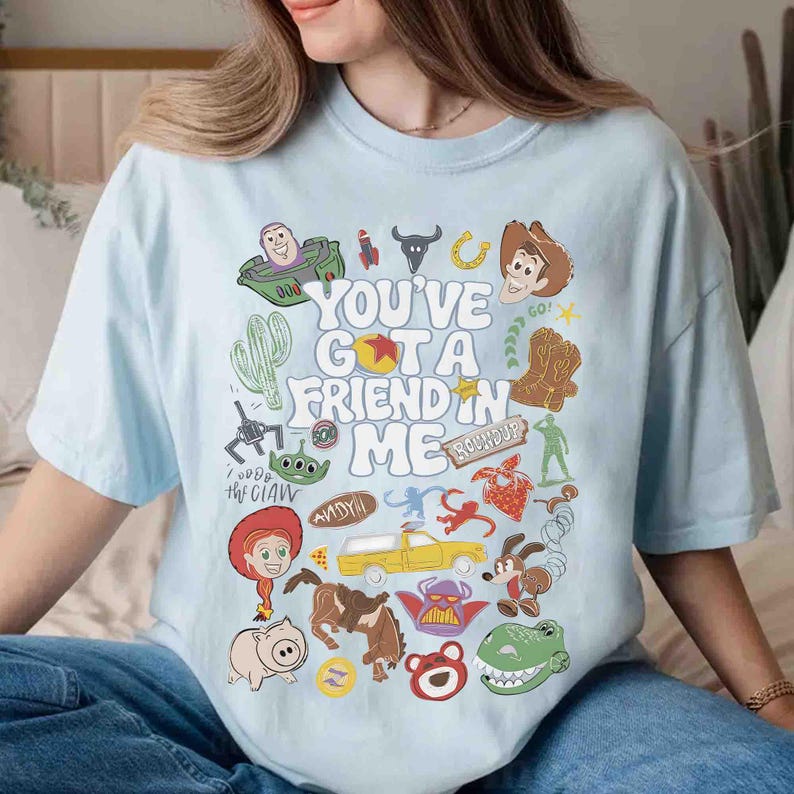 May include: Light blue t-shirt with the phrase "You've Got a Friend in Me" and various cartoon character graphics. The design includes a cactus, a rocket, a horseshoe, a cowboy, and a pig.