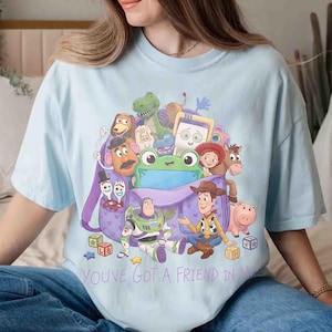 May include: A light blue t-shirt featuring a graphic print of various animated characters from Toy Story emerging from a purple backpack. The design includes Buzz Lightyear, Woody, and other figures, with the text YOU'VE GOT A FRIEND IN ME printed below the illustration.