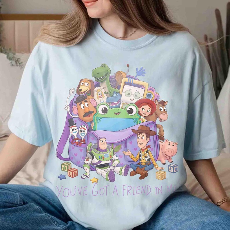May include: A light blue t-shirt featuring a graphic print of various animated characters from Toy Story emerging from a purple backpack. The design includes Buzz Lightyear, Woody, and other figures, with the text YOU'VE GOT A FRIEND IN ME printed below the illustration.