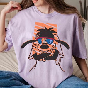 May include: A lavender t-shirt with a cartoon image of a dog wearing sunglasses. The dog is wearing a red and blue bandana and has a white and orange background.