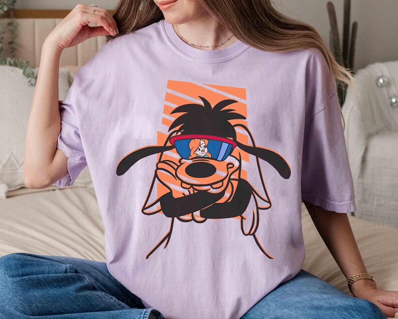 May include: A lavender t-shirt with a cartoon image of a dog wearing sunglasses. The dog is wearing a red and blue bandana and has a white and orange background.