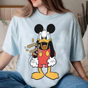 May include: Pale blue t-shirt with a cartoon Donald Duck design. The character is depicted in a red outfit, yellow feet, and a black body. Speech bubbles with the word "QUACK" and stars are also included.