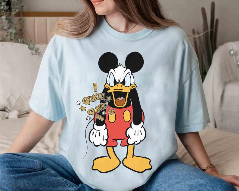May include: Pale blue t-shirt with a cartoon Donald Duck design. The character is depicted in a red outfit, yellow feet, and a black body. Speech bubbles with the word "QUACK" and stars are also included.
